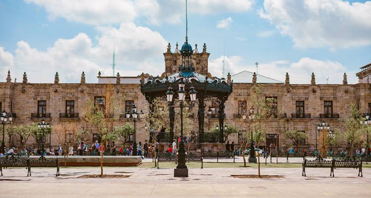 Plaza histórica con quiosco de música ornamentado de hierro forjado, edificio gubernamental de piedra y familias paseando bajo nubes esponjosas.