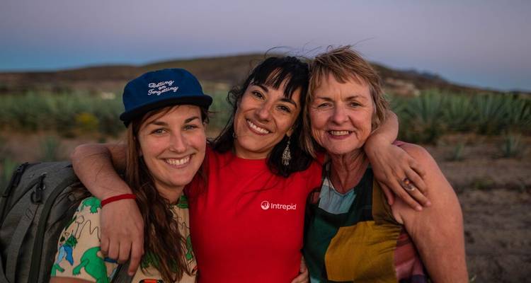 Tres mujeres sonriendo con los brazos alrededor una de la otra en un paisaje desértico de agaves al atardecer.