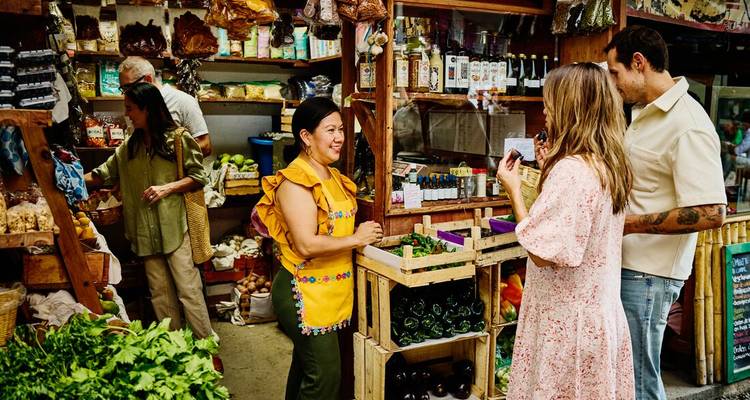 Puesto vibrante de mercado mexicano donde los viajeros conversan con un vendedor local rodeado de productos frescos.