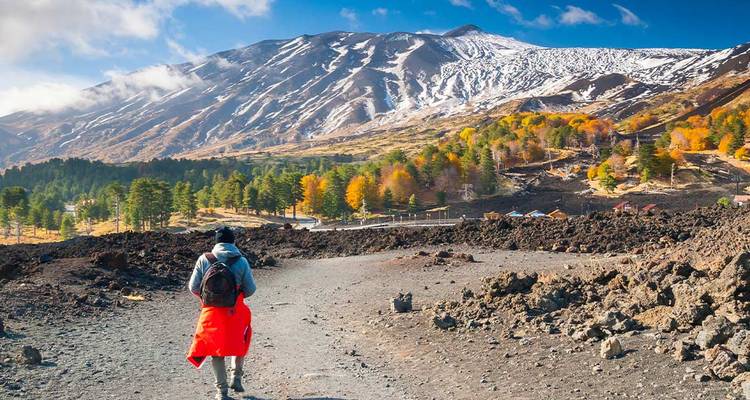 Excursionista con chaqueta roja se acerca a las escarpadas laderas volcánicas del Monte Etna cubierto de nieve bajo un cielo azul brillante.