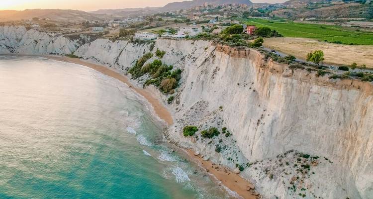 Vista aérea de acantilados blancos resplandecientes y mar turquesa acariciando una playa siciliana apartada al atardecer.