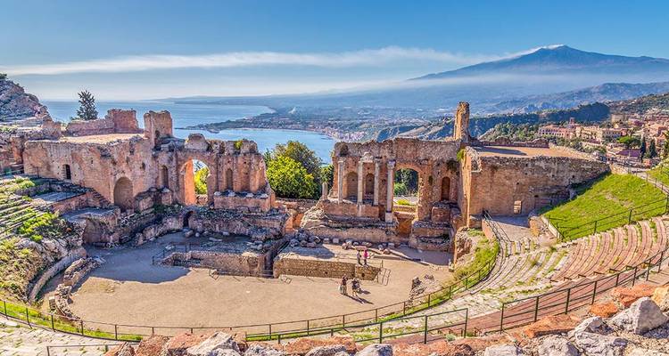 Las ruinas del teatro griego antiguo en Taormina contemplan una bahía resplandeciente con el Monte Etna humeando en la distancia.