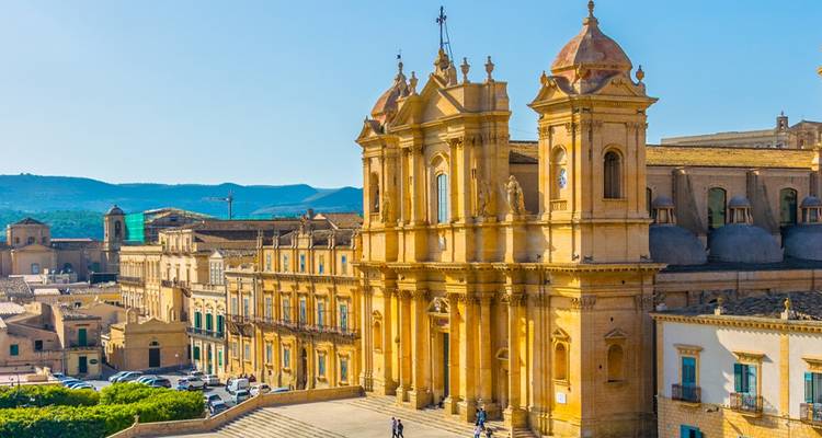 La fachada barroca dorada de la Catedral de Noto resplandece bajo la luz de la tarde sobre una gran escalinata y la plaza del casco antiguo.