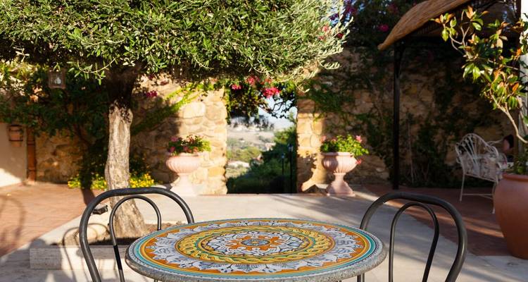 Patio soleado con una mesa de azulejos de mosaico bajo un olivo con vista a las colinas rurales sicilianas.