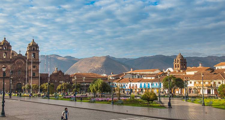 Weite Aussicht auf Cuscos Plaza de Armas mit prächtigen Kathedralen, Gärten und den fernen Anden unter gemusterten Wolken.