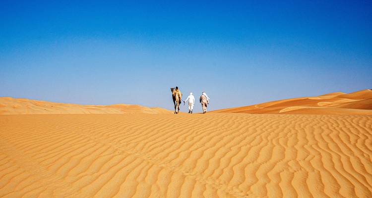 Twee mannen in traditionele kledij die een kameel over eindeloze golvende zandduinen onder een onberispelijke blauwe lucht leiden.