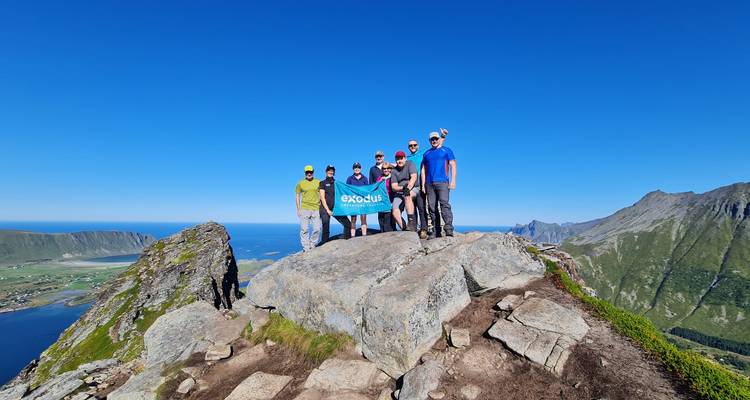 Un pequeño grupo de senderismo se para en una cumbre rocosa con vistas panorámicas del mar y las montañas bajo un cielo azul despejado.