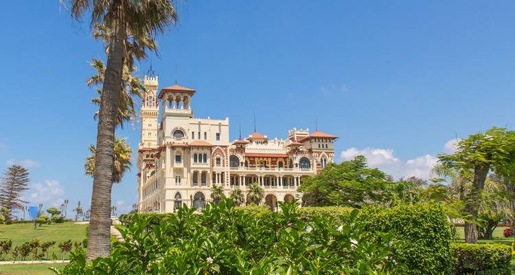 Montaza Palace steekt uit boven weelderig groen en palmbomen onder een strakblauwe Alexandrijnse lucht.