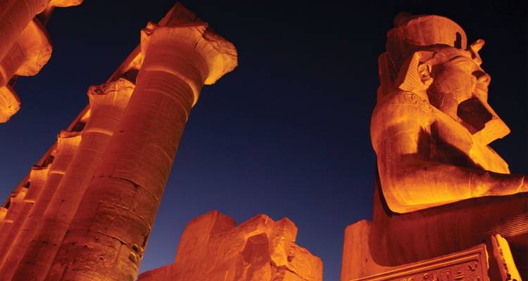 Columnas antiguas iluminadas y una estatua de un faraón sentado brillan en color naranja contra un cielo azul oscuro y despejado.