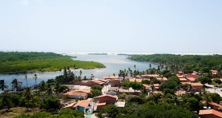 Village côtier avec des toits en terre cuite au bord d'une rivière bordée de mangroves s'ouvrant sur des dunes de sable clair et l'horizon océanique.