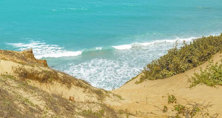 Steile zandige helling met schaarse begroeiing die afloopt naar turquoise oceaangolven.