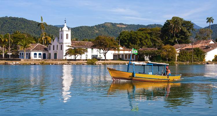 Una colorida embarcación de pesca pasa frente al malecón colonial de Paraty con una iglesia blanca y colinas boscosas detrás.