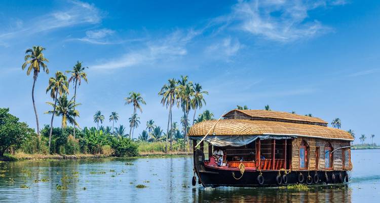 Ein traditionelles hölzernes Hausboot gleitet entlang der Backwaters von Kerala, die von Kokospalmen gesäumt sind, unter einem klaren blauen Himmel.
