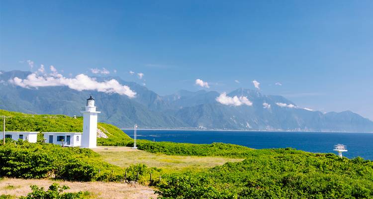 Faro en un acantilado costero con hierba y montañas al fondo.