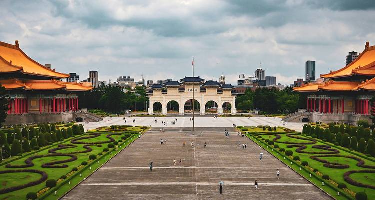 Gran plaza con edificios ornamentados y jardines, horizonte de la ciudad al fondo.