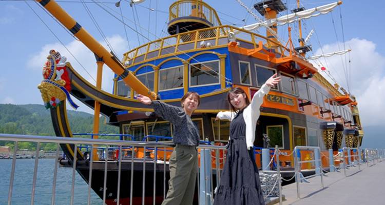 Twee vrouwen nemen speelse poses aan op een pier naast een kleurrijk piraten-sightseeing schip.