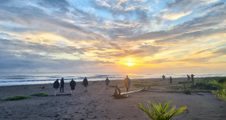 Un grupo de viaje camina por una playa de arena negra al amanecer con un cielo colorido que se refleja en las olas.