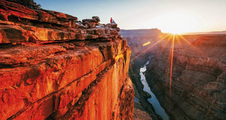 Personne assise au bord d'une falaise au coucher du soleil, surplombant un canyon.