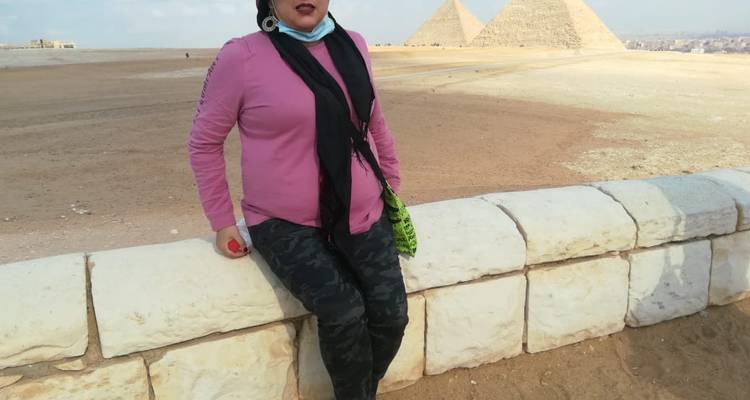 Een vrouw die op een stenen hek zit met de piramides op de achtergrond.