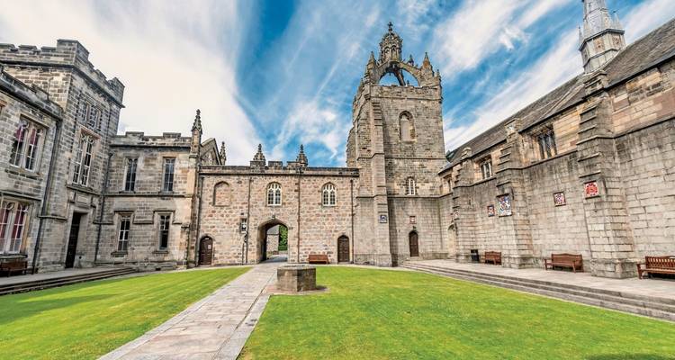 A courtyard surrounded by stone buildings with a towering structure in the center and a grassy lawn.