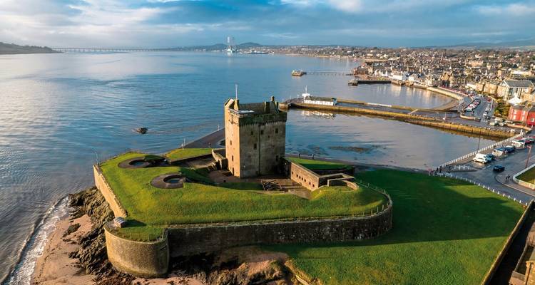 Aerial view of a coastal fortification with a central tower, surrounded by water and a bridge in the distance.