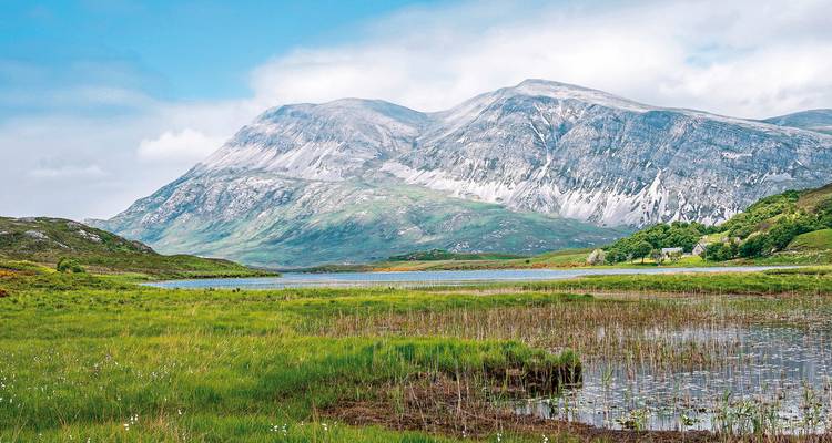 A scenic view of a mountainous landscape with green fields and a lake in the foreground.