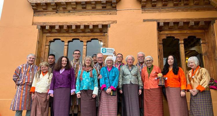 Grupo de turistas vistiendo ropa tradicional bhutanesa posando felices frente a un edificio color ocre.