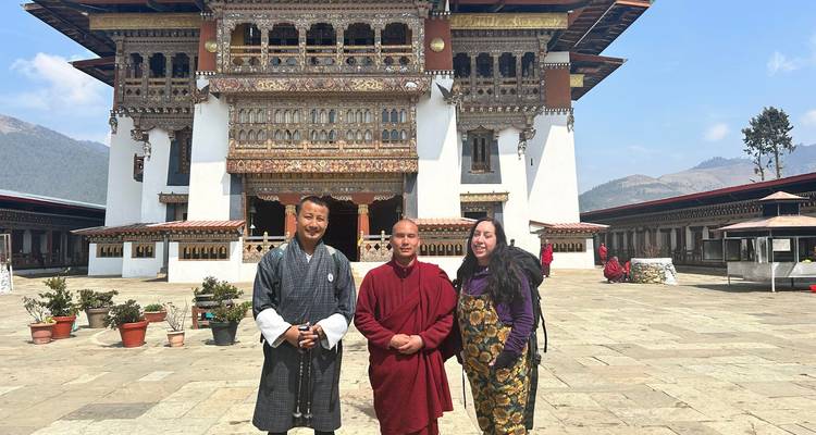 Dos visitantes y un monje sonríen frente a un patio de dzong butanés elaboradamente decorado.