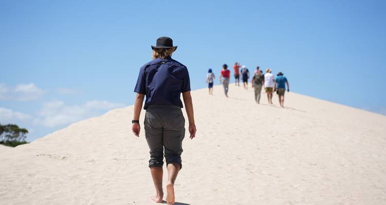 Guía descalzo lidera un pequeño grupo subiendo una duna de arena blanca empinada bajo un cielo azul brillante