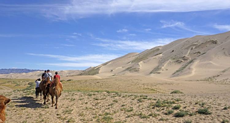 People riding camels across sand dunes under a blue sky.