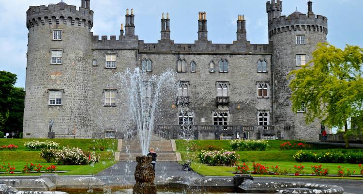 Château historique en pierre avec de hautes tours rondes derrière une fontaine éclatante et des jardins entretenus.