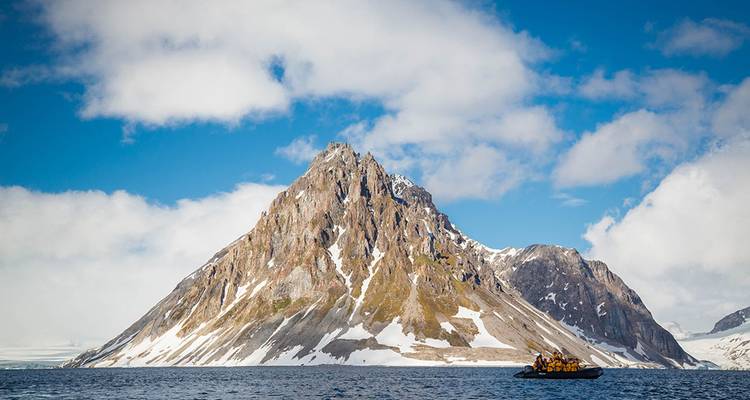Une montagne rugueuse striée de neige s'élève abruptement des eaux arctiques tandis qu'un petit Zodiac rempli de voyageurs emmitouflés s'approche.