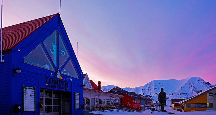 Bâtiment bleu coloré du centre commercial Lompen avec des montagnes enneigées qui brillent dans un ciel de coucher de soleil violet en arrière-plan.