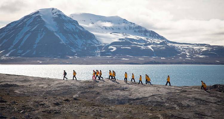 Une procession de voyageurs en vestes jaunes traverse un terrain rocheux austère au bord d'une baie glaciaire avec de hautes montagnes enneigées au-delà.