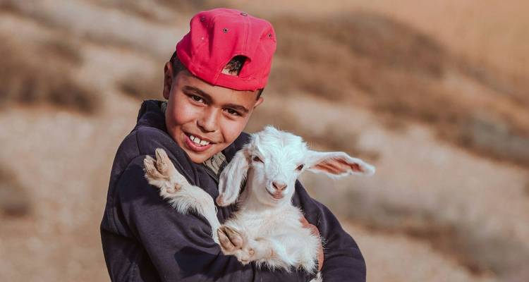 Een jongen die een geit vasthoudt met een woestijnlandschap op de achtergrond.