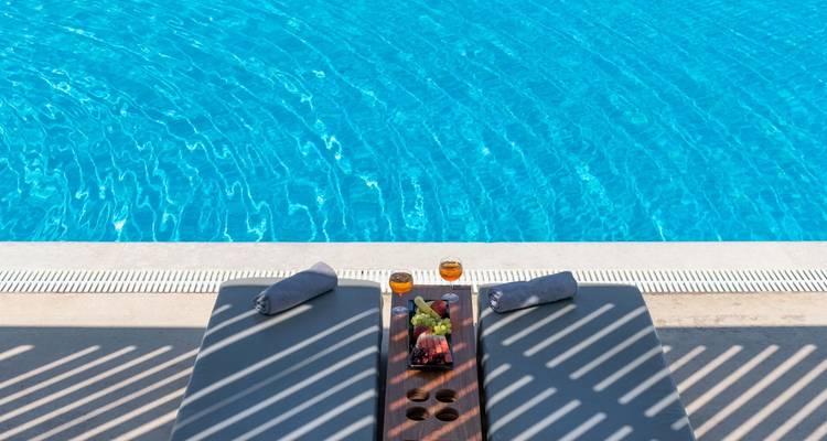 Des chaises longues au bord de la piscine avec un plateau de fruits et des serviettes à côté d'une eau bleue cristalline.