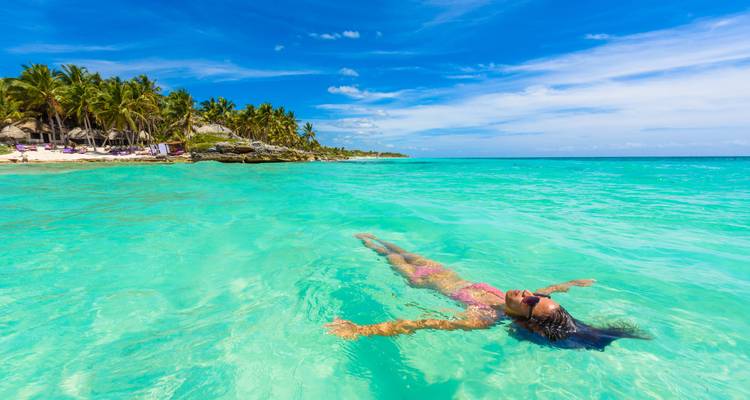 Persoon drijvend in helder turquoise water nabij een zandstrand met palmbomen.