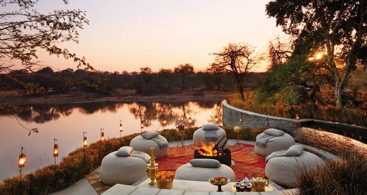 Außensitzbereich an einem See mit Feuerstelle und Laternen, aufgenommen bei Sonnenuntergang.
