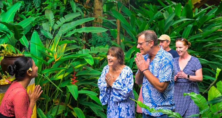 Balinese host greets smiling visitors with traditional gesture amid lush tropical garden.