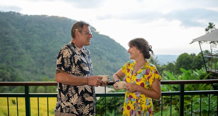 Mature couple enjoys coffee on a balcony overlooking forested hills.