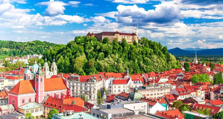 Ein Bergschloss krönt eine üppig grüne Stadt mit rotgedeckten Gebäuden unter einem strahlend blauen Himmel.
