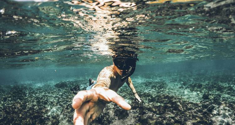 Persona haciendo snórkel bajo el agua, extendiendo la mano hacia la cámara.