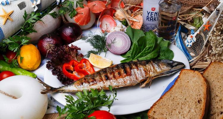 Plato de pescado a la parrilla completo decorado con verduras frescas, pan y hierbas al lado de un vaso de raki