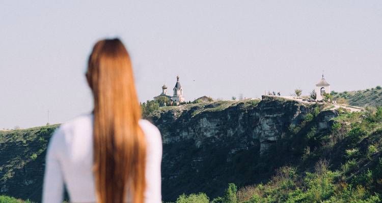 Out-of-focus woman looking toward cliff-edge monastery under clear sky.