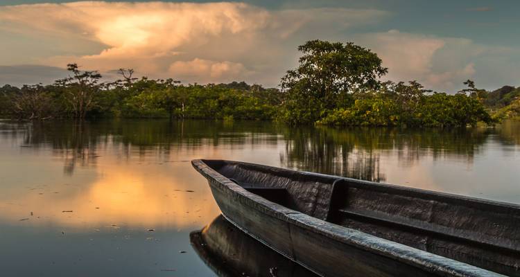 Houten uitgeholde kano drijvend op kalm spiegelglad water bij zonsondergang met jungle bomen daarachter