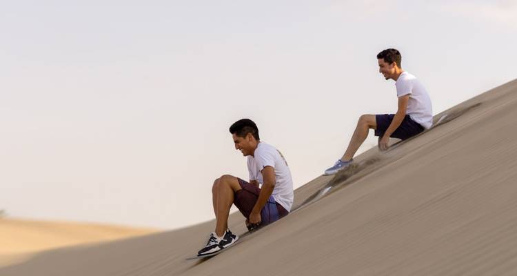 Zwei Personen fahren Sandbrett einen Düne hinunter.