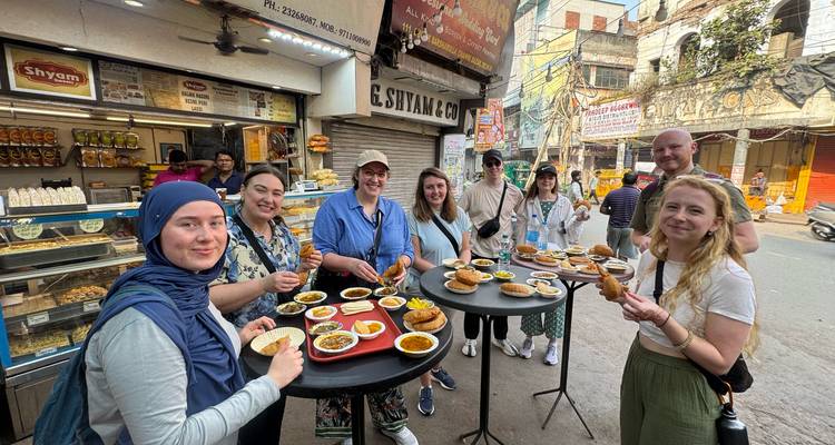 Touristen probieren eine Vielzahl von indischen Straßensnacks an hohen Tischen außerhalb einer Bäckerei.