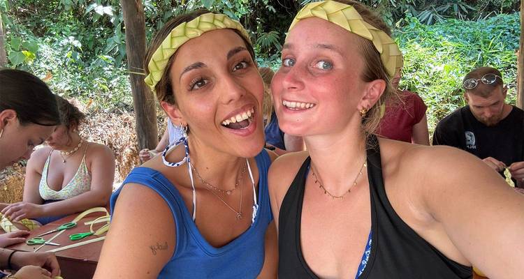 Deux femmes souriantes prennent un autoportrait rapproché en portant des bandeaux faits à la main en feuilles de palmier lors d'un atelier d'artisanat.