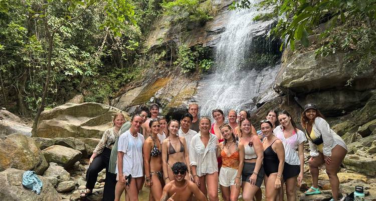 Un grand groupe de touristes en maillot de bain posant à la base d'une cascade tropicale entourée de rochers et de forêt.