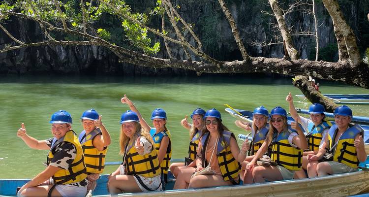 Lächelnde Gruppe in Rettungswesten und blauen Helmen, die Daumen hoch zeigen, während sie in Ruderbooten auf einem grünen Fluss unterhalb von Kalksteinfelsen sitzen.